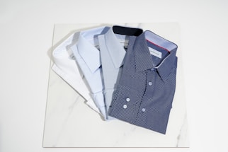 Four neatly folded dress shirts are arranged on a light-colored marble surface. The shirts vary in color and pattern, including white, light blue, and two shades of blue with small dot patterns. The collars and buttons of the shirts are clearly visible.