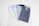 Four neatly folded dress shirts are arranged on a light-colored marble surface. The shirts vary in color and pattern, including white, light blue, and two shades of blue with small dot patterns. The collars and buttons of the shirts are clearly visible.