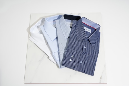 Four neatly folded dress shirts are arranged on a light-colored marble surface. The shirts vary in color and pattern, including white, light blue, and two shades of blue with small dot patterns. The collars and buttons of the shirts are clearly visible.
