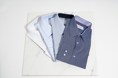Four neatly folded dress shirts are arranged on a light-colored marble surface. The shirts vary in color and pattern, including white, light blue, and two shades of blue with small dot patterns. The collars and buttons of the shirts are clearly visible.