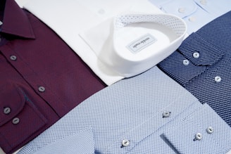 Several neatly arranged dress shirts featuring various patterns and colors, including white, burgundy, dark blue, and light blue. The shirts display button details and fine fabric textures, reflecting a sense of organization and style.