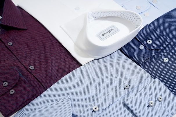 Several neatly arranged dress shirts featuring various patterns and colors, including white, burgundy, dark blue, and light blue. The shirts display button details and fine fabric textures, reflecting a sense of organization and style.