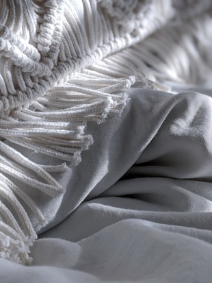 Close-up of a textured blanket or fabric with intricate woven patterns and fringes on a smooth surface, showing soft folds and subtle shadows.