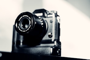 A black camera with a large, prominent lens is set against a soft, blurred background. The camera appears vintage or classic in design, with metallic details and a textured body.