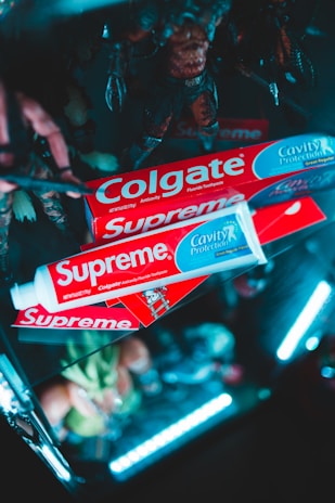 Several Colgate toothpaste boxes are creatively arranged with bright lighting. The packaging is branded with both Colgate and Supreme logos. The background includes blurred figurines and a dark setting with neon lighting for a striking effect.