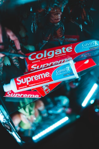 Several Colgate toothpaste boxes are creatively arranged with bright lighting. The packaging is branded with both Colgate and Supreme logos. The background includes blurred figurines and a dark setting with neon lighting for a striking effect.