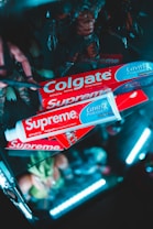 Several Colgate toothpaste boxes are creatively arranged with bright lighting. The packaging is branded with both Colgate and Supreme logos. The background includes blurred figurines and a dark setting with neon lighting for a striking effect.
