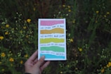 A hand holding a colorful book with Spanish text over a background of green foliage and yellow flowers. The book cover features a series of horizontal colored stripes.