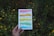 A hand holding a colorful book with Spanish text over a background of green foliage and yellow flowers. The book cover features a series of horizontal colored stripes.