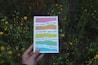 A hand holding a colorful book with Spanish text over a background of green foliage and yellow flowers. The book cover features a series of horizontal colored stripes.