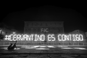 A black and white nighttime scene with a large illuminated sign that reads '#GERMANYINISE CONTIGO'. The sign is constructed on a metal frame with bright lights outlining each letter. In the foreground, two people are sitting and observing the sign. The background includes a large building with a faintly visible facade and windows.