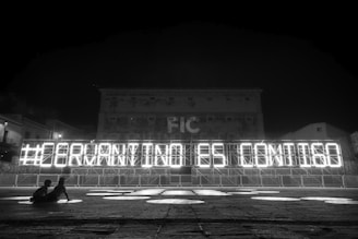 A black and white nighttime scene with a large illuminated sign that reads '#GERMANYINISE CONTIGO'. The sign is constructed on a metal frame with bright lights outlining each letter. In the foreground, two people are sitting and observing the sign. The background includes a large building with a faintly visible facade and windows.