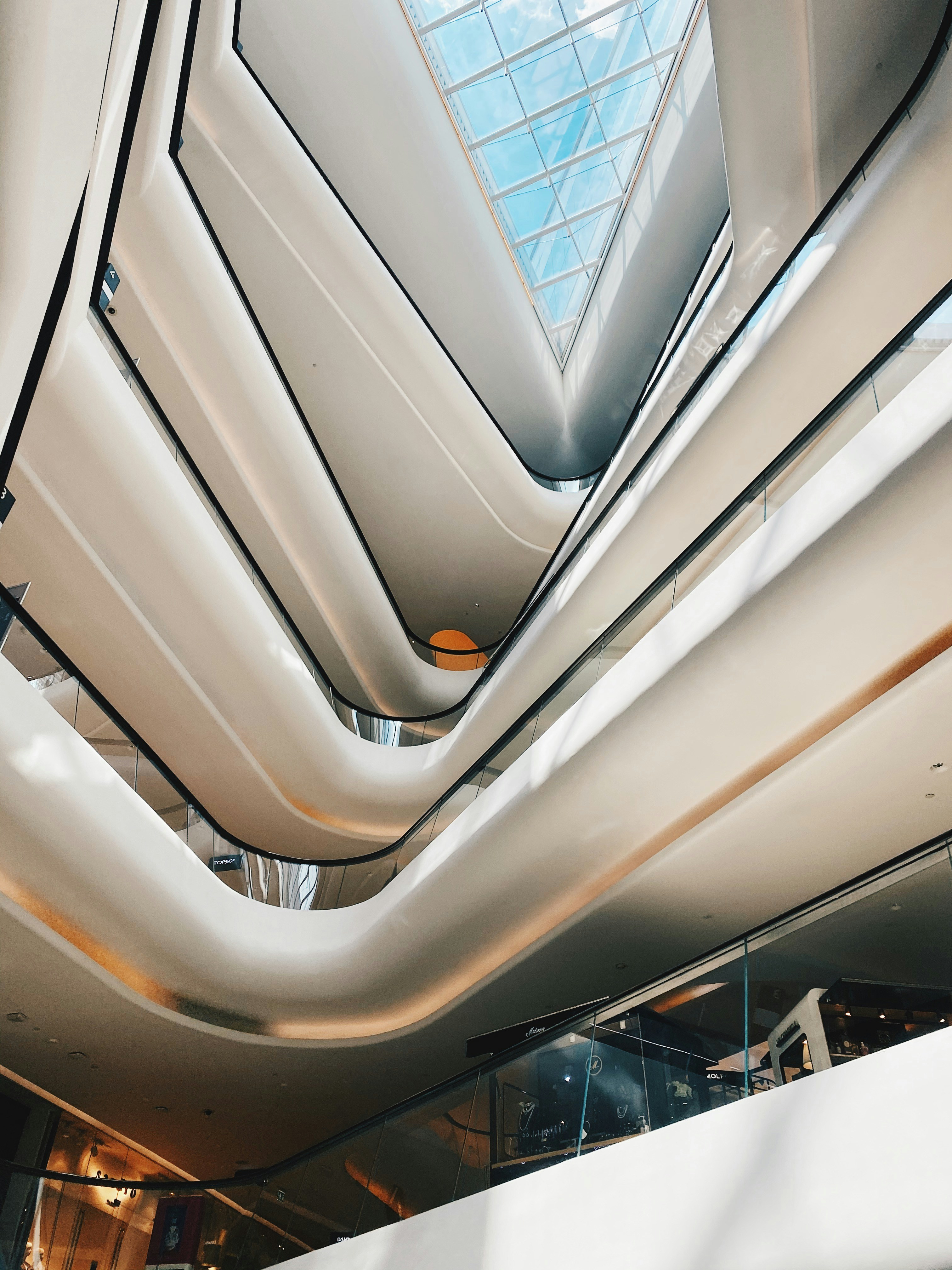 An architectural interior with multiple floors, featuring sleek, modern design elements and abundant natural light from a large glass ceiling. The structure showcases flowing lines and a bright, open space.