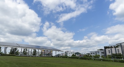 A grassy field under a partly cloudy sky. On the left, there is a row of solar panels elevated on a structure. Buildings are visible in the background with a mix of greenery and urban architecture.