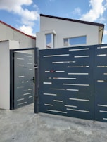 A modern house with a flat roof and large rectangular windows is located behind a futuristic gate with a sleek, perforated design. The gate is slightly open, situated on a concrete driveway. The sky is partly cloudy, contributing to a serene residential scene.