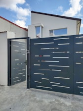 A modern house with a flat roof and large rectangular windows is located behind a futuristic gate with a sleek, perforated design. The gate is slightly open, situated on a concrete driveway. The sky is partly cloudy, contributing to a serene residential scene.