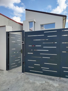 A modern house with a flat roof and large rectangular windows is located behind a futuristic gate with a sleek, perforated design. The gate is slightly open, situated on a concrete driveway. The sky is partly cloudy, contributing to a serene residential scene.