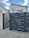 A modern house with a flat roof and large rectangular windows is located behind a futuristic gate with a sleek, perforated design. The gate is slightly open, situated on a concrete driveway. The sky is partly cloudy, contributing to a serene residential scene.
