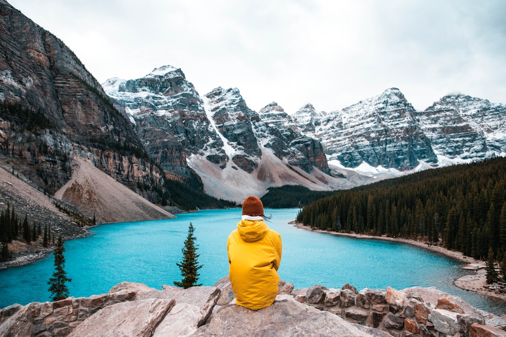 pessoa em capuz amarelo sentado na rocha perto do lago e montanha coberta de neve durante o dia