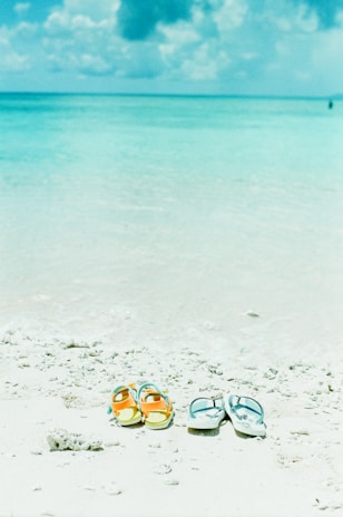 Two pairs of sandals sit on a bright, sandy beach with turquoise water in the background under a sky with fluffy clouds. The scene conveys a sense of relaxation and leisure.