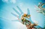 A vivid, multicolored dragon is depicted against a bright blue sky. The dragon, intricately detailed with green scales, orange accents, and flowing whiskers, is layered with the shadow of a hand, creating a surreal and artistic effect. Wispy clouds add texture to the sky, enhancing the ethereal quality of the image.