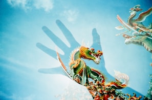 A vivid, multicolored dragon is depicted against a bright blue sky. The dragon, intricately detailed with green scales, orange accents, and flowing whiskers, is layered with the shadow of a hand, creating a surreal and artistic effect. Wispy clouds add texture to the sky, enhancing the ethereal quality of the image.