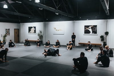 A group of people wearing martial arts uniforms are sitting on a large mat in a spacious room with high ceilings. The walls are adorned with art pieces and there are several potted plants placed around the room. The scene conveys a martial arts training session or class.