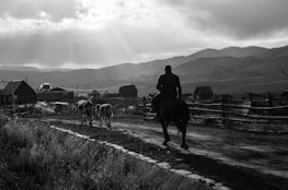 A rural scene features a man riding a horse down a dirt path, followed by two cows. The landscape is mountainous, with sunlight filtering through clouds, casting dramatic shadows. Wooden fences and modest houses populate the background, enhancing the rustic atmosphere.