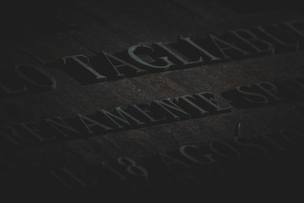 Engraved text on a stone surface, partially illuminated and casting deep shadows, with a somber and aged appearance. There is a presence of moss or discoloration on the stone.