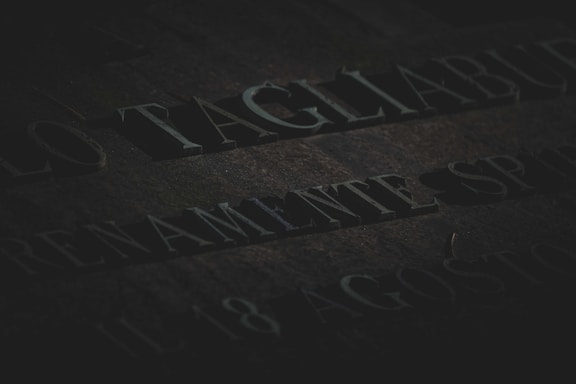 Engraved text on a stone surface, partially illuminated and casting deep shadows, with a somber and aged appearance. There is a presence of moss or discoloration on the stone.
