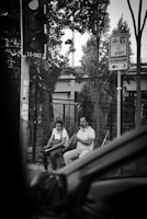 Two individuals are sitting on a street, with one playing a wind instrument and the other sitting beside with an accordion. They seem to be performing near a fence with urban structures in the background. A traffic light and street signs are visible, and there is a partial view of a vehicle in the foreground.