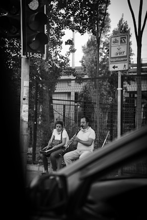 Two individuals are sitting on a street, with one playing a wind instrument and the other sitting beside with an accordion. They seem to be performing near a fence with urban structures in the background. A traffic light and street signs are visible, and there is a partial view of a vehicle in the foreground.