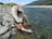 An angler is kneeling by a clear mountain river, holding a fishing net with a large fish inside. The surrounding landscape features rocky banks and distant green hills under a partly cloudy sky.