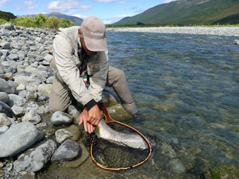 An angler is kneeling by a clear mountain river, holding a fishing net with a large fish inside. The surrounding landscape features rocky banks and distant green hills under a partly cloudy sky.