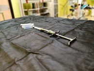 A syringe is placed on a dark cloth-covered surface alongside several vials. The background is slightly blurred, indicating a laboratory or medical setting, with shelves and equipment visible.