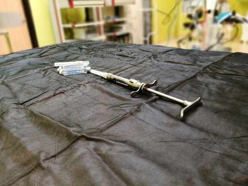 A syringe is placed on a dark cloth-covered surface alongside several vials. The background is slightly blurred, indicating a laboratory or medical setting, with shelves and equipment visible.