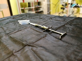 A syringe is placed on a dark cloth-covered surface alongside several vials. The background is slightly blurred, indicating a laboratory or medical setting, with shelves and equipment visible.