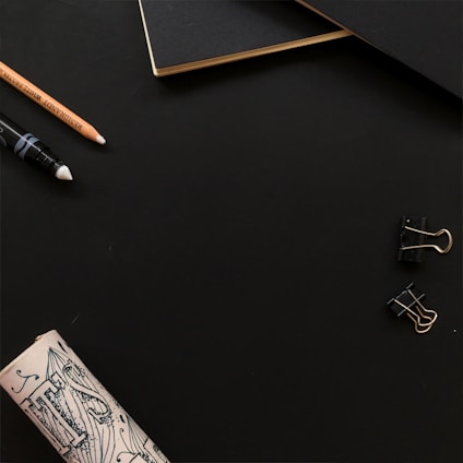 A minimalist workspace setup featuring a black notebook with gold-edged pages, a pencil, a white-tipped drawing pen, two black binder clips, and a rolled-up piece of illustrated paper against a dark background.
