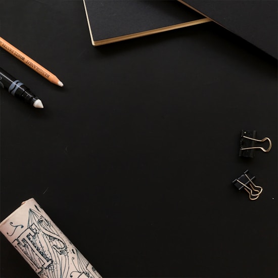A minimalist workspace setup featuring a black notebook with gold-edged pages, a pencil, a white-tipped drawing pen, two black binder clips, and a rolled-up piece of illustrated paper against a dark background.