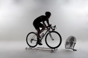 A person is cycling on an indoor trainer bike with a fan positioned in front. The setting appears minimalistic with a plain background, and the cyclist is wearing cycling gear, including a helmet.