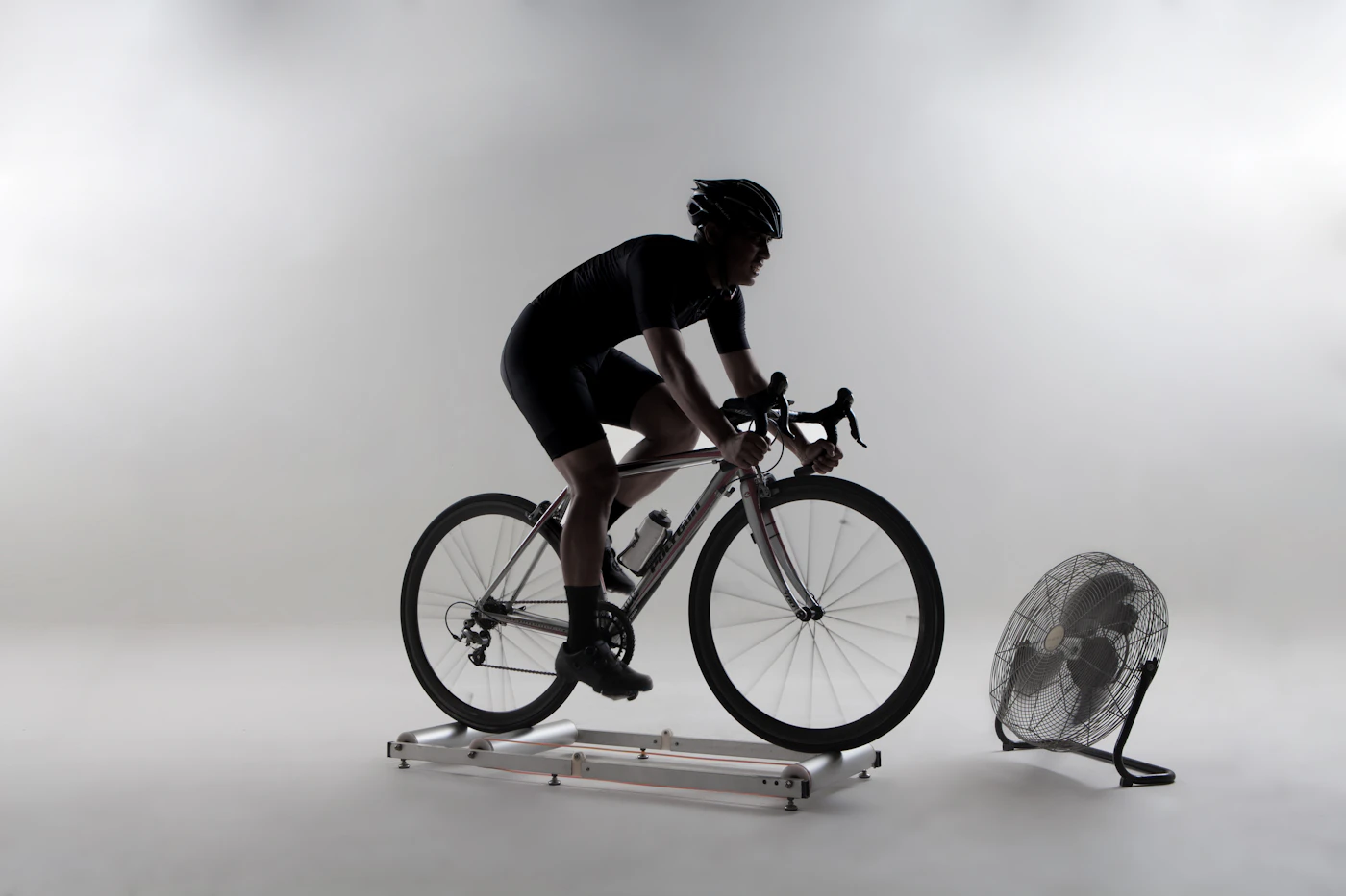 A cyclist riding rollers indoors with a fan alongside