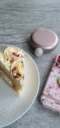 A slice of cake with creamy frosting and decorative toppings sits on a patterned white plate. Nearby is a rose gold compact case and a phone with a floral design cover resting on a stone-textured surface.