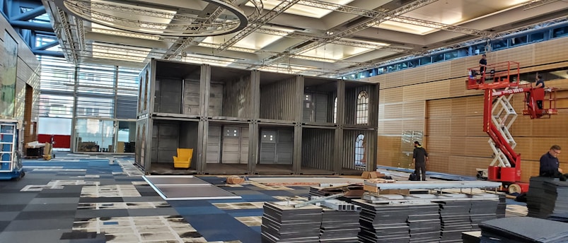 A large indoor space with a structure made of stacked shipping containers, one of which has a yellow chair inside. The area is being set up with several workers visible, one on a raised platform. The space is well-lit with a modern ceiling and large windows.