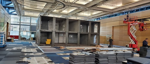 A large indoor space with a structure made of stacked shipping containers, one of which has a yellow chair inside. The area is being set up with several workers visible, one on a raised platform. The space is well-lit with a modern ceiling and large windows.