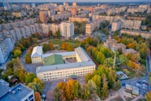 An aerial view of a cityscape showcasing a school complex with a U-shaped building in the foreground. The structure is surrounded by lush greenery with trees exhibiting various autumn colors. In the background, there are numerous residential high-rise buildings stretching across the horizon.