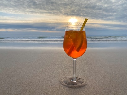 A glass of orange-colored drink with ice and a straw is placed on the sand near a beach. The ocean waves and the horizon are visible in the background beneath a partly cloudy sky, with the sun setting or rising.