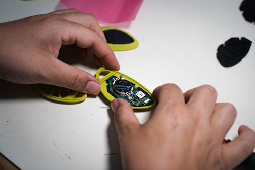 A pair of hands is handling a small electronic device, which is open to reveal its internal components, including a circuit board and a lithium button cell battery labeled CR2032. The device case is yellow and is being held on a white surface with some other parts scattered around.