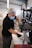 A person wearing a mask and gloves is working on assembling or repairing a metal structure, possibly a bicycle or motorcycle, in a workshop setting with various tools and equipment visible in the background.