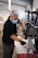 A person wearing a mask and gloves is working on assembling or repairing a metal structure, possibly a bicycle or motorcycle, in a workshop setting with various tools and equipment visible in the background.