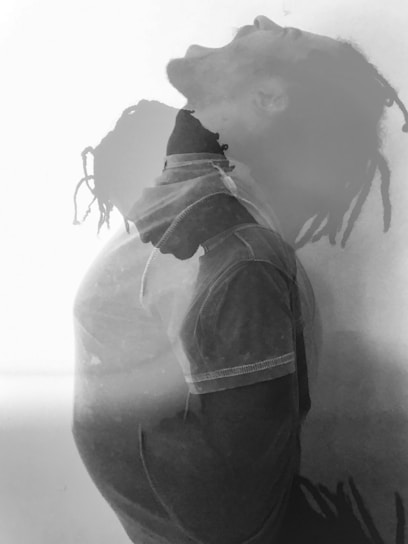 A double exposure effect features a person captured in two overlapping poses. One pose shows an upward, open-mouthed gesture, possibly expressing an intense emotion, while the other presents a reflective stance looking down. The composition is monochromatic, emphasizing shadows and light contrasts.
